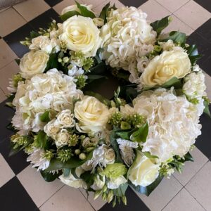 Composition de fleurs piquées dans la mousse synthétique imbibée d’eau à la forme circulaire appelée : couronne.Modele présenté en taille S en camaïeu de blanc et vert